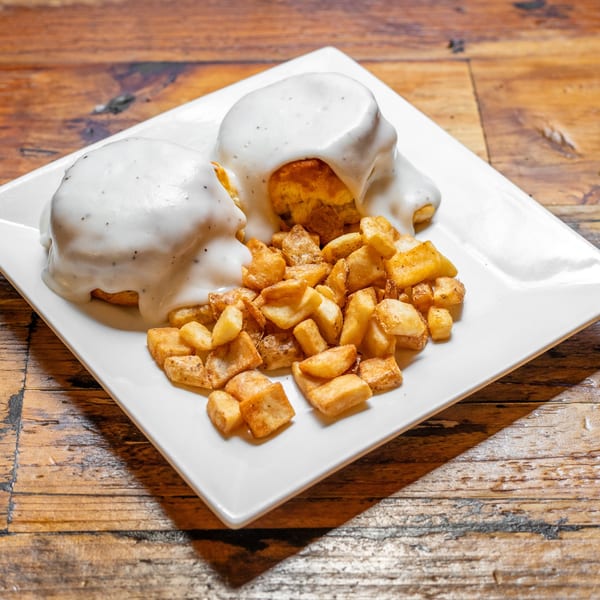 Two biscuits covered in white gravy with black pepper, served with a side of crispy home fries.
