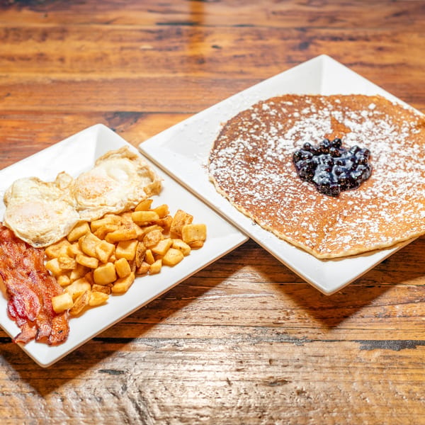 A breakfast platter with bacon, home fries, fried eggs, and a pancake with blueberries.