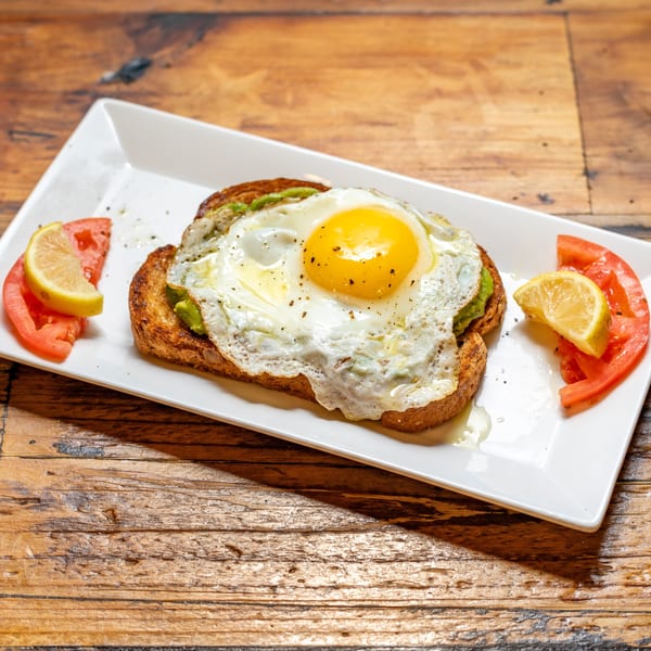 Avocado toast with a fried egg, garnished with tomato slices and lemon wedges on a white plate.